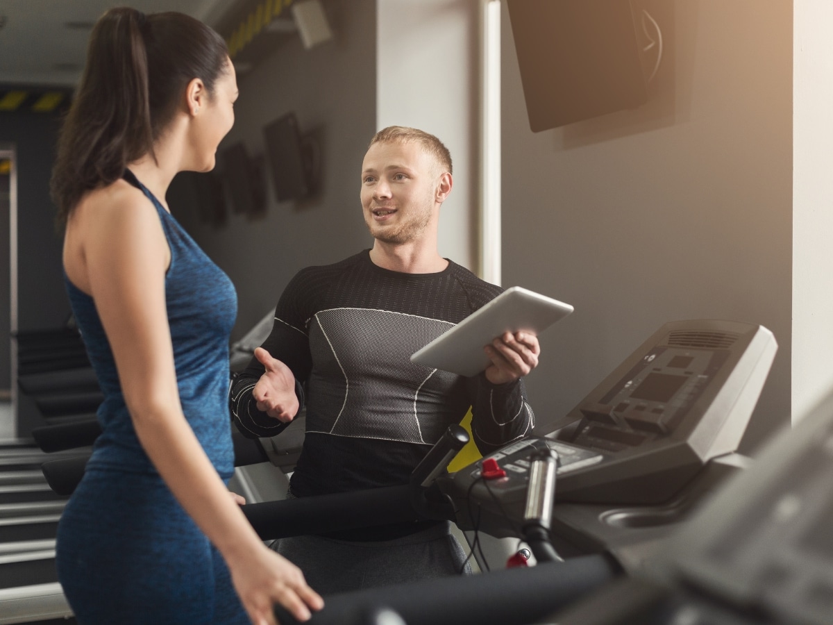 Fitness instructor helps young woman on elliptical trainer. Coach discussing workout plan with girl on treadmill. Cardio workout in gym, healthy lifestyle.